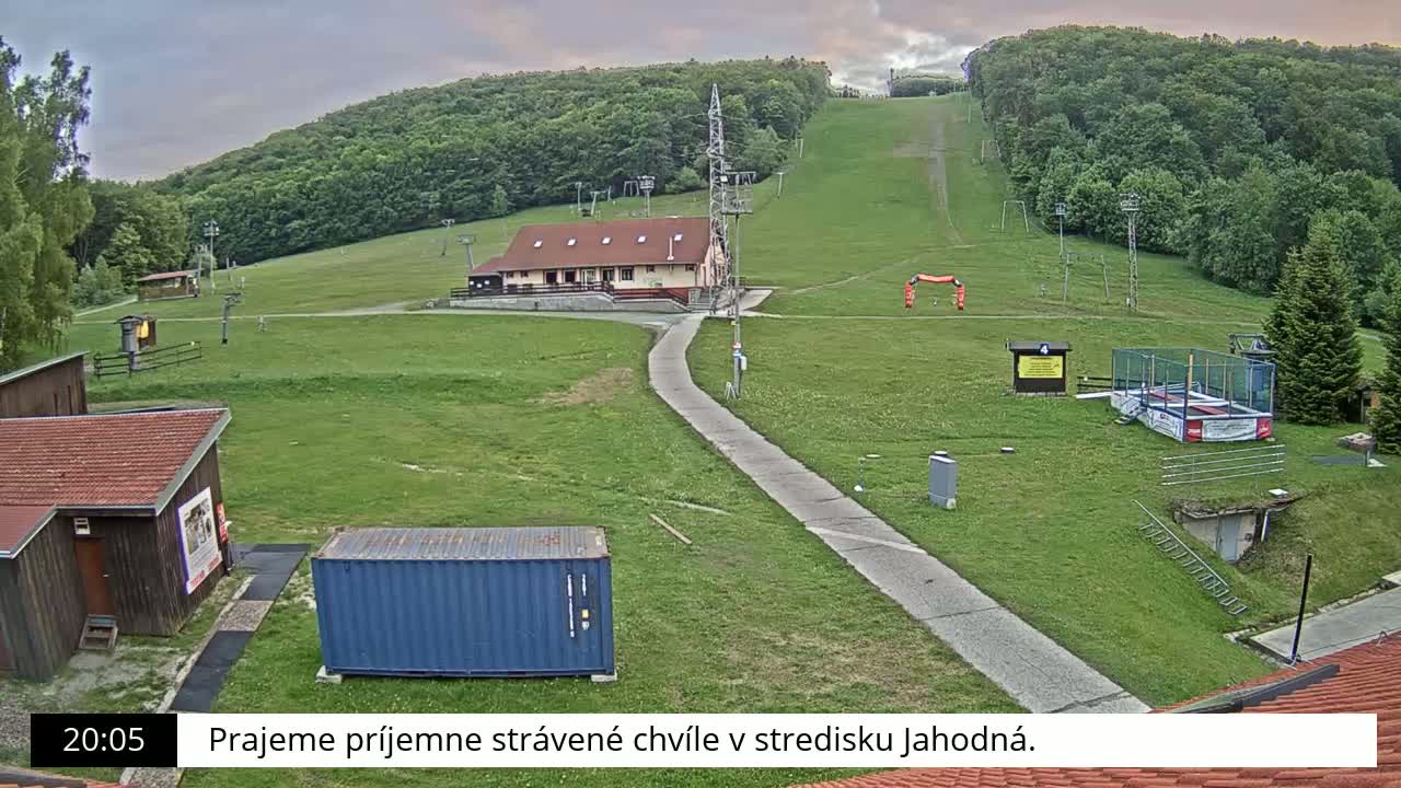 A ski slope with a building, several smaller structures, and a trampoline sits on a grassy hillside under an overcast sky.