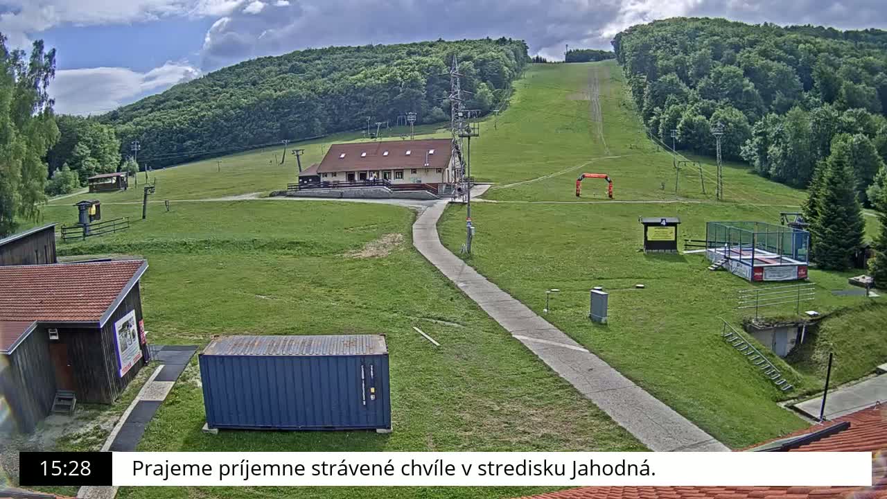 A grassy ski slope with a building, a paved path, and a shipping container is visible under a partly cloudy sky.