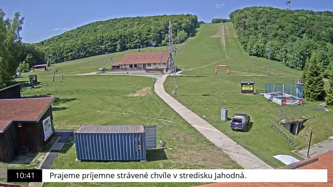 A ski slope with a building, grassy areas, and a few vehicles is visible under a sunny sky.