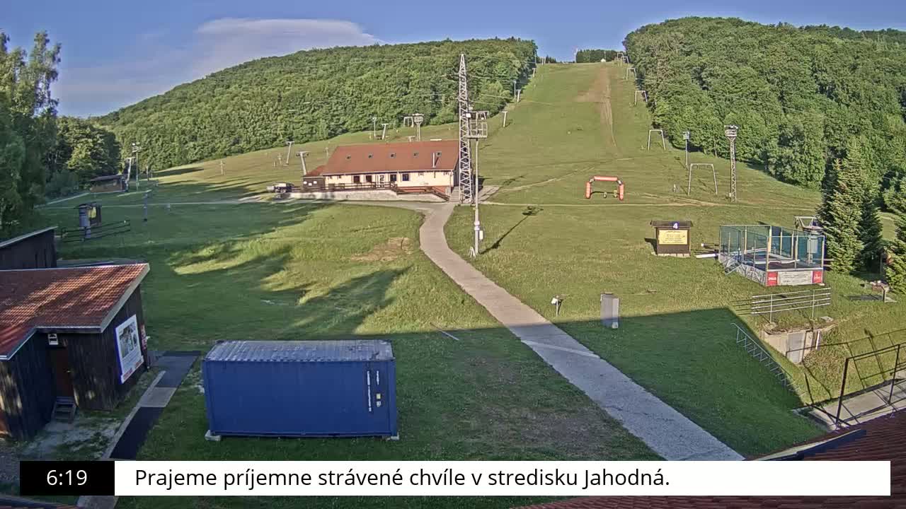 A grassy ski slope with a building and several smaller structures is visible under a mostly sunny sky.