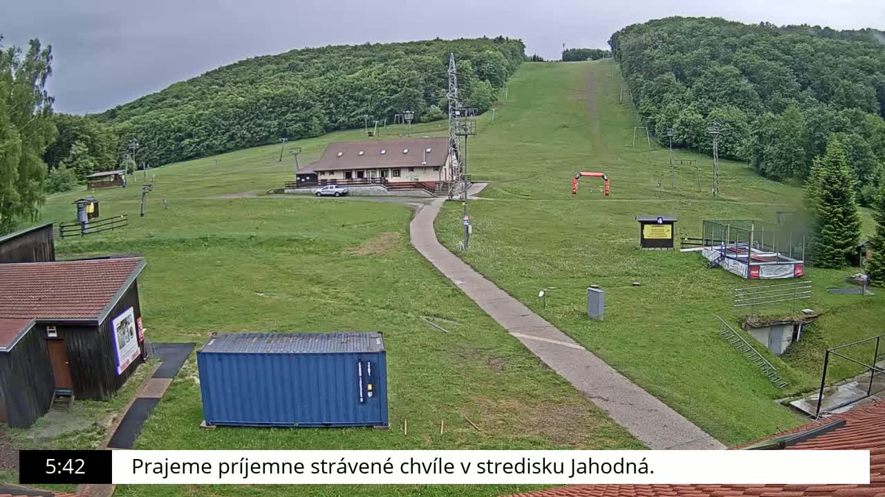 A grassy ski slope with a building, a paved path, and overcast skies.
