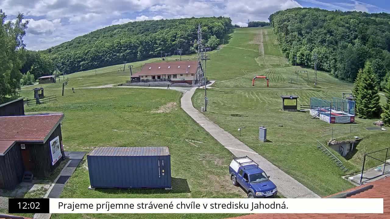 A grassy ski slope with a small building, a pickup truck, and a shipping container is visible under a partly cloudy sky.