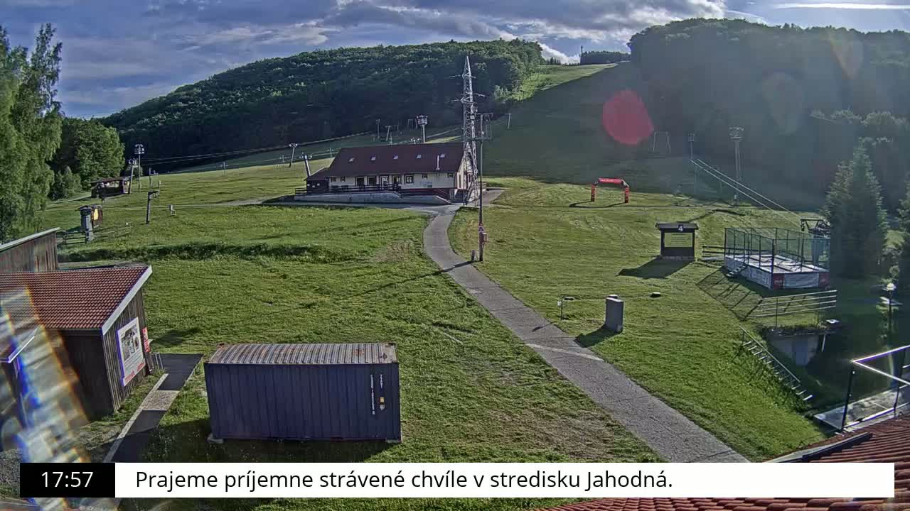 A grassy ski slope with a building, chairlifts, and other structures, under a partly cloudy sky.