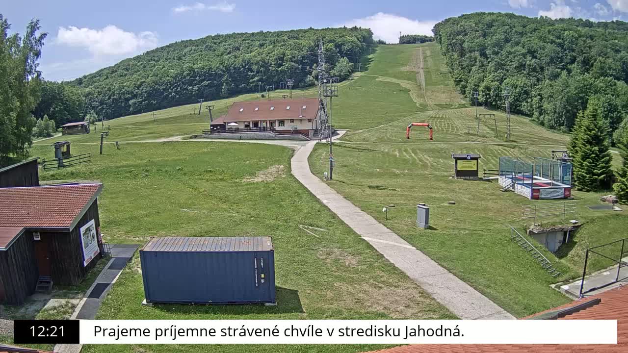 A grassy hillside with a building, ski lift infrastructure, and a paved path, under sunny skies.