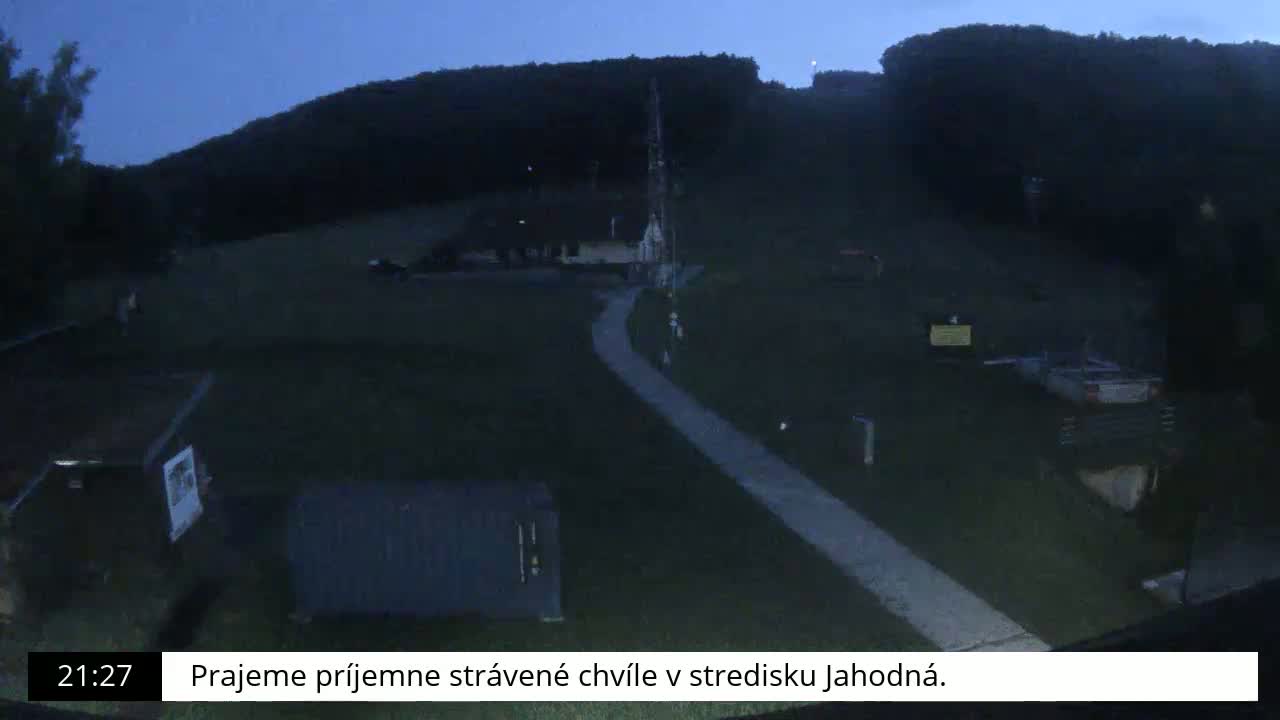 A paved pathway winds through a dark, grassy area toward a building at the base of a hill under a twilight sky.