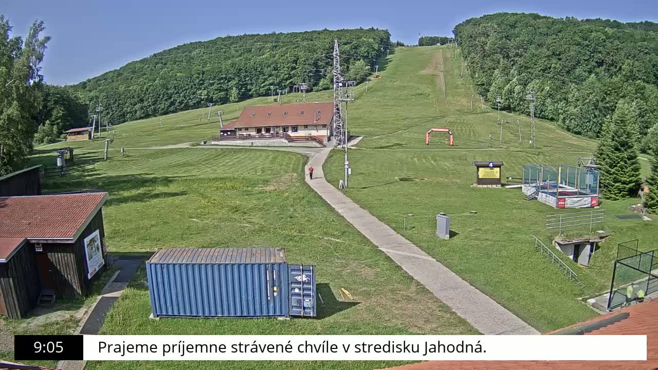 A grassy hillside with a ski lift, small buildings, and a person walking on a path on a sunny day.