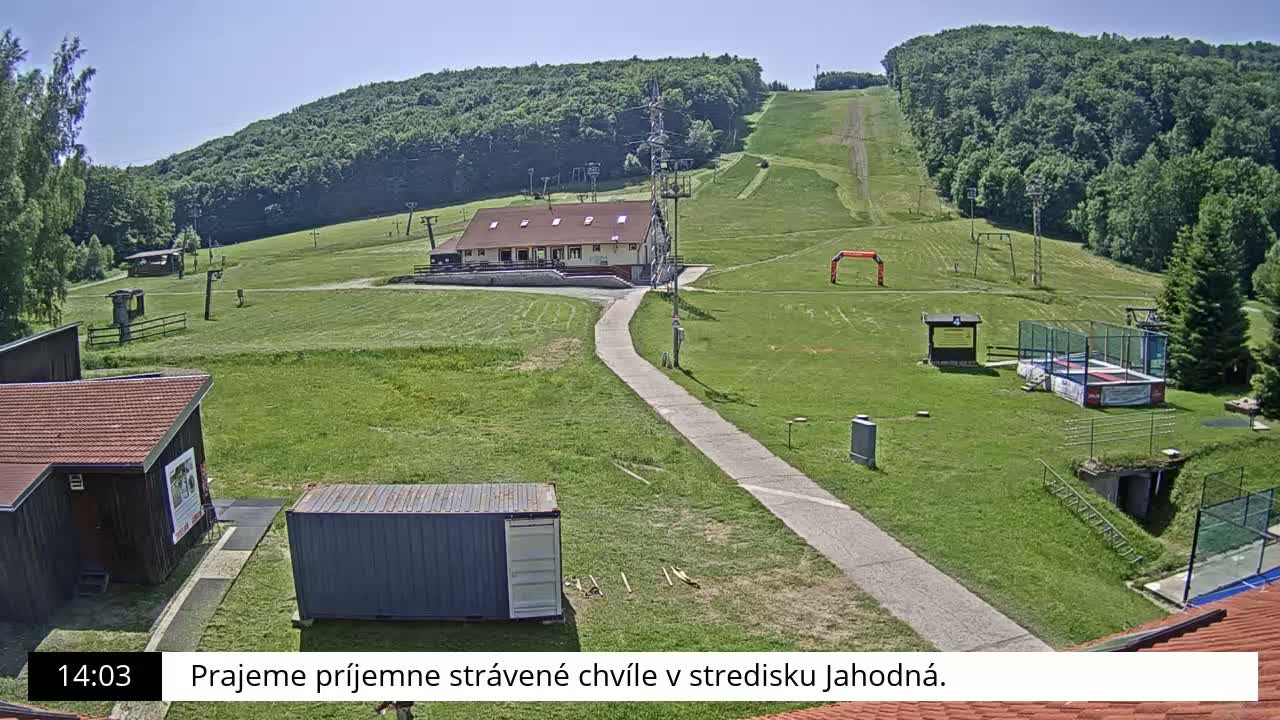 A grassy ski slope with a building, small structures, and a paved path is visible on a sunny day.