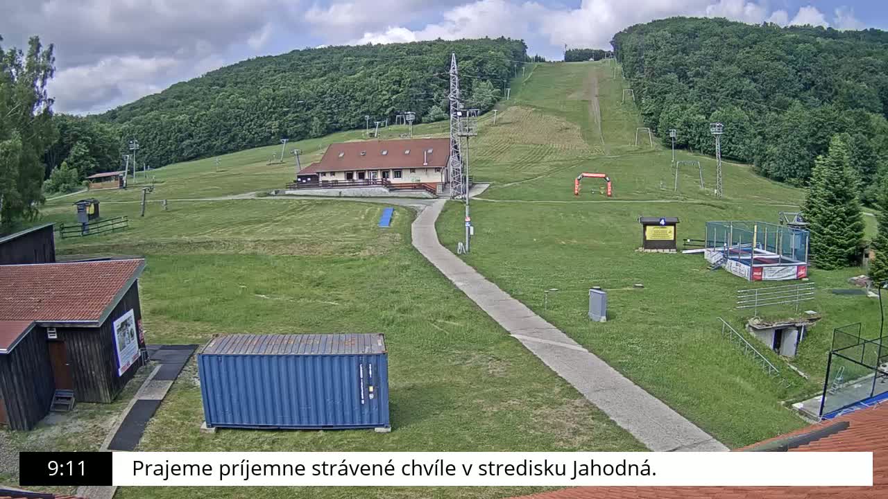A grassy hill with a ski lift, a building, and other recreational facilities is visible under a partly cloudy sky.