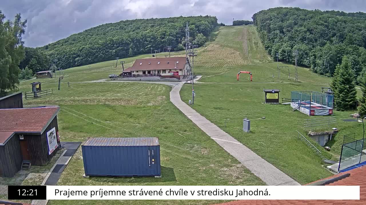 A grassy ski slope with a building, a paved path, and some recreational equipment is visible under a partly cloudy sky.