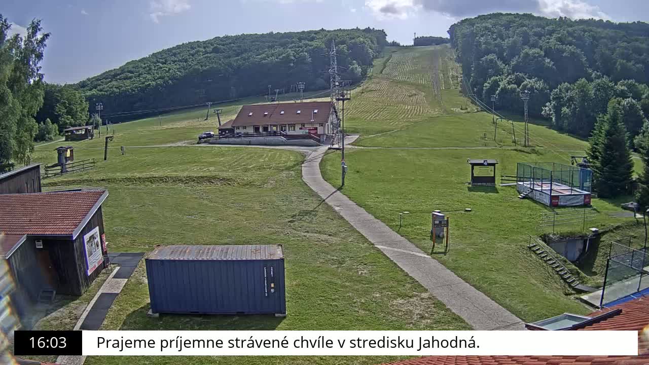 A grassy hillside with a ski lift, a small building, and a paved pathway is visible under a partly sunny sky.