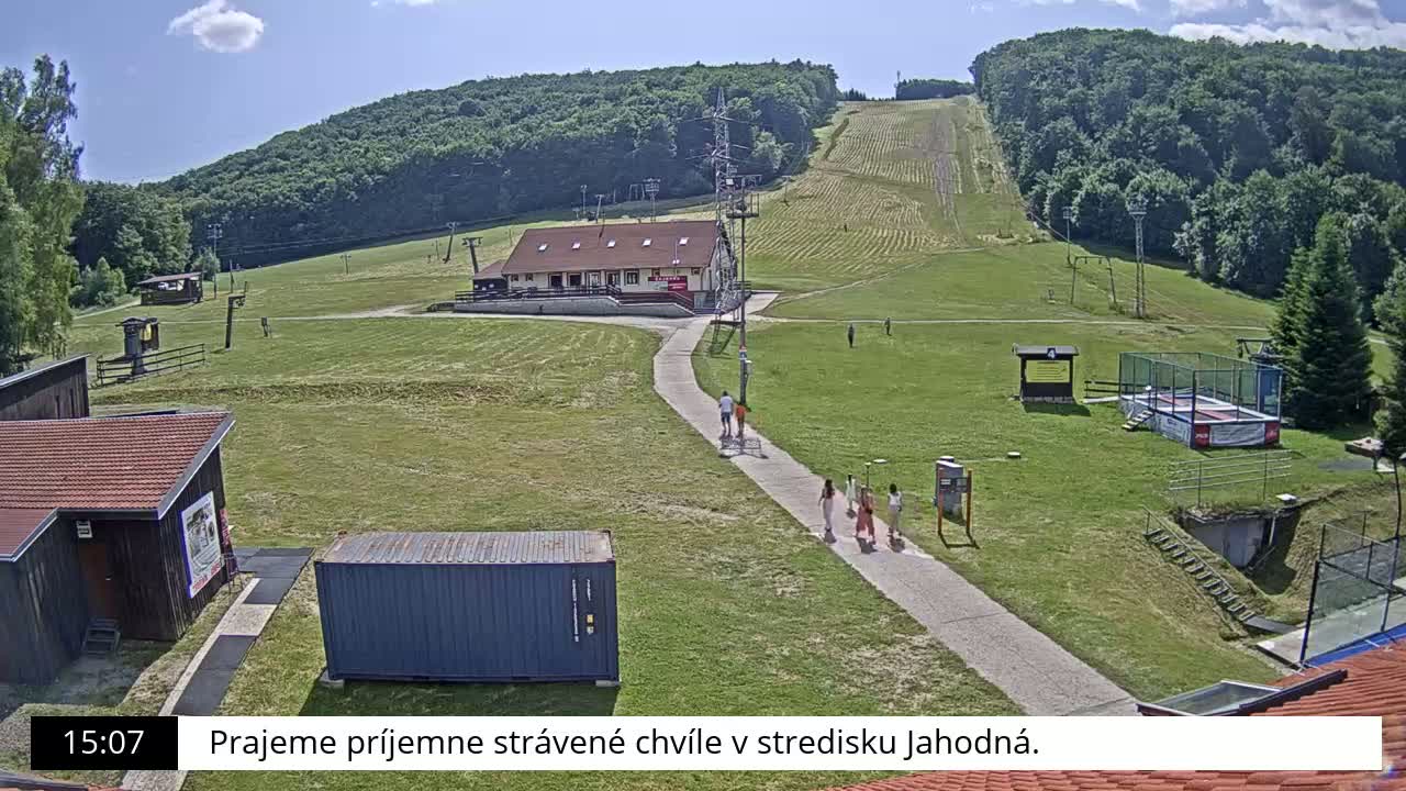 A grassy hillside with a ski lift and several buildings, including a small lodge, is visible on a sunny day, with several people walking along a path.