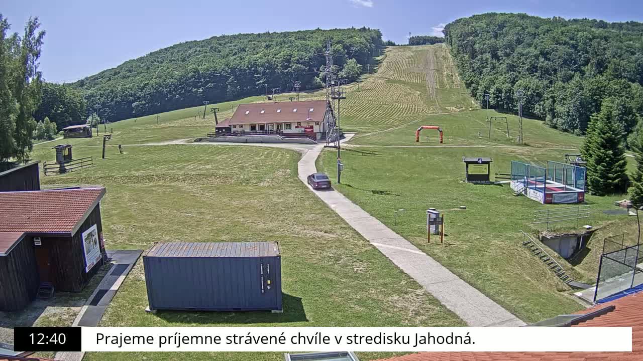 A sunny day at a ski resort with a grassy area, a building, a car, and ski lift infrastructure is visible.