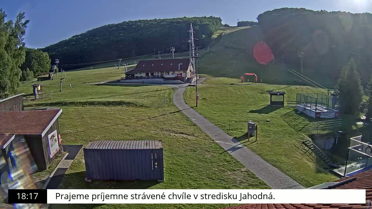 A grassy ski resort with a building, lift towers, and a paved pathway is visible on a sunny day.