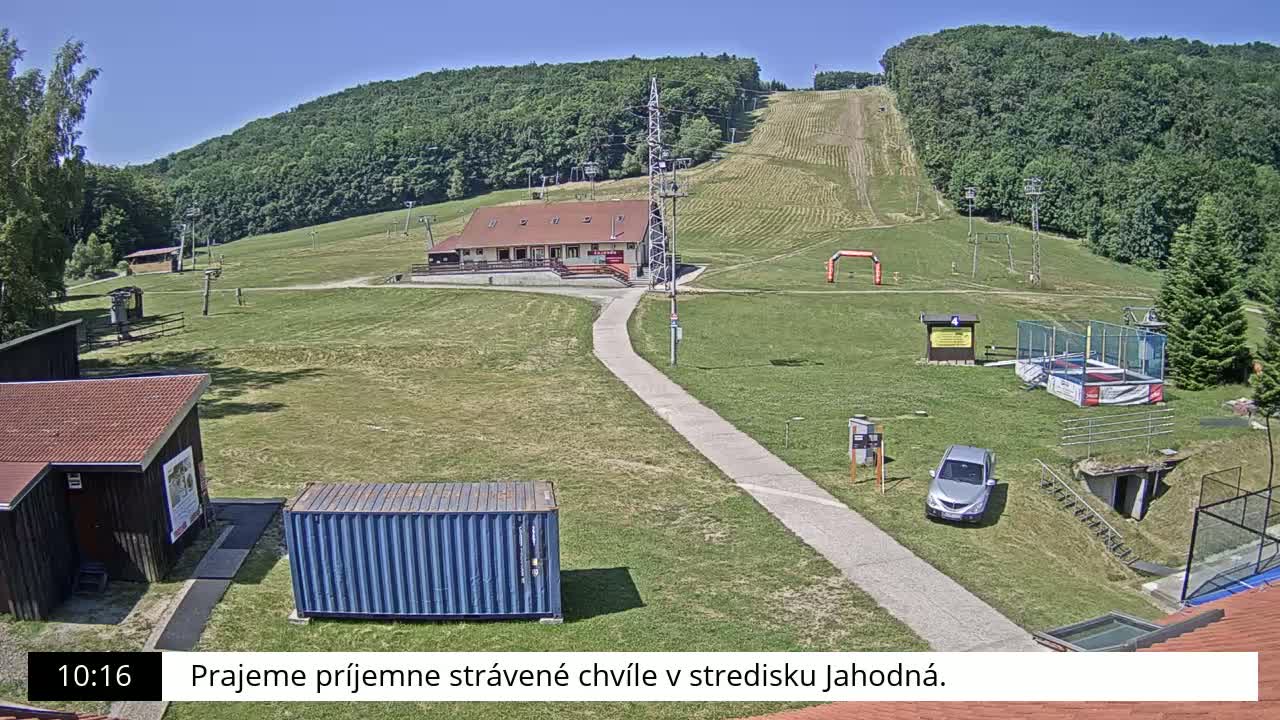 A grassy ski slope with a building, a car, and other recreational facilities is visible on a sunny day.