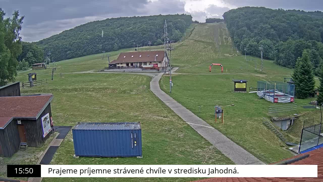 A grassy ski slope with a building, several smaller structures, and a trampoline is visible under an overcast sky.