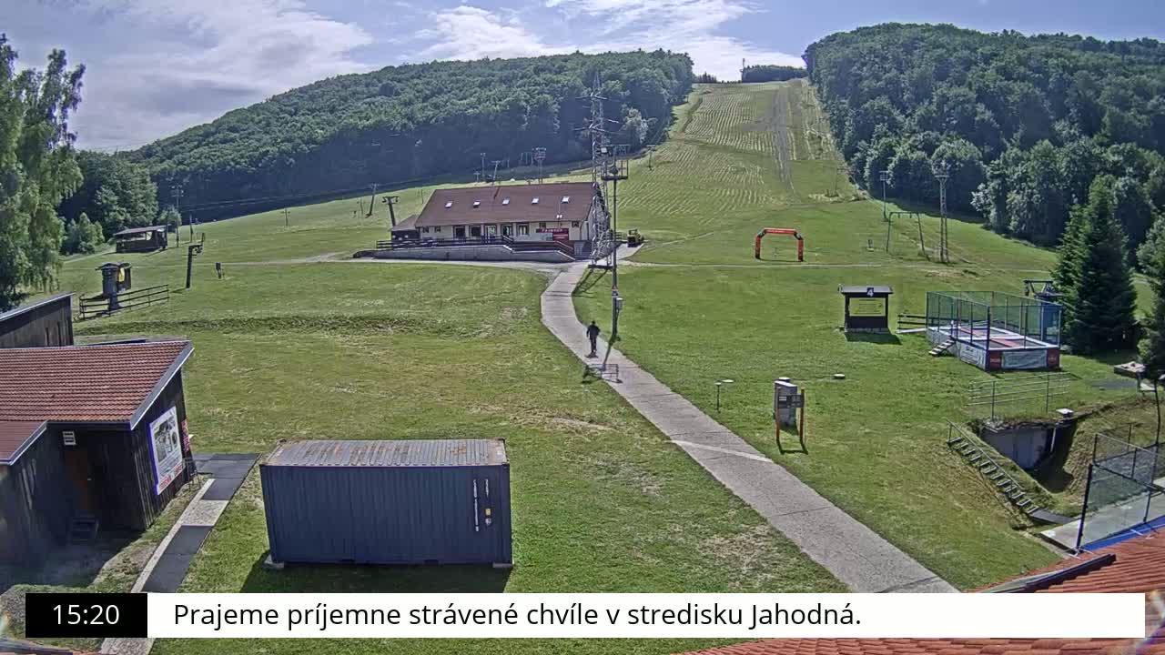 A grassy hillside with a ski lift, small buildings, a walking path, and a person walking, all under sunny skies.