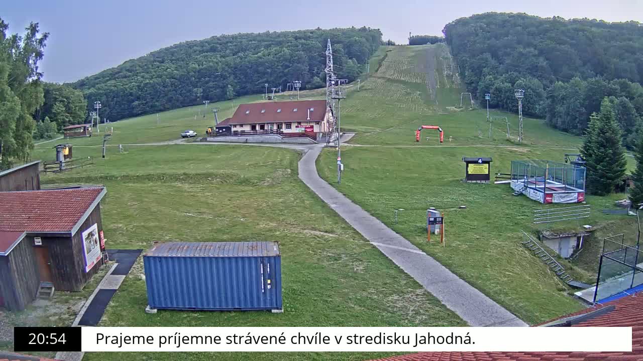 A grassy ski slope with a building, several small structures, and a blue shipping container is visible under a clear sky.