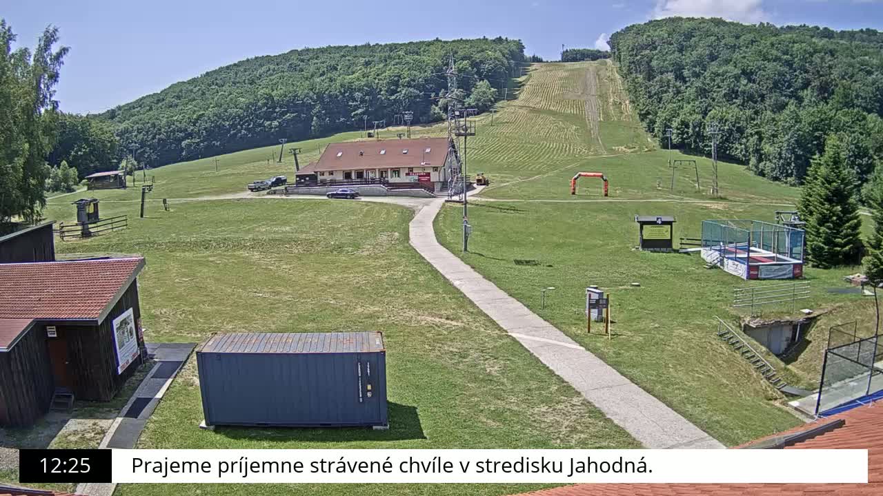 A grassy hillside ski resort with a building, a paved path, and various recreational facilities is visible on a sunny day.