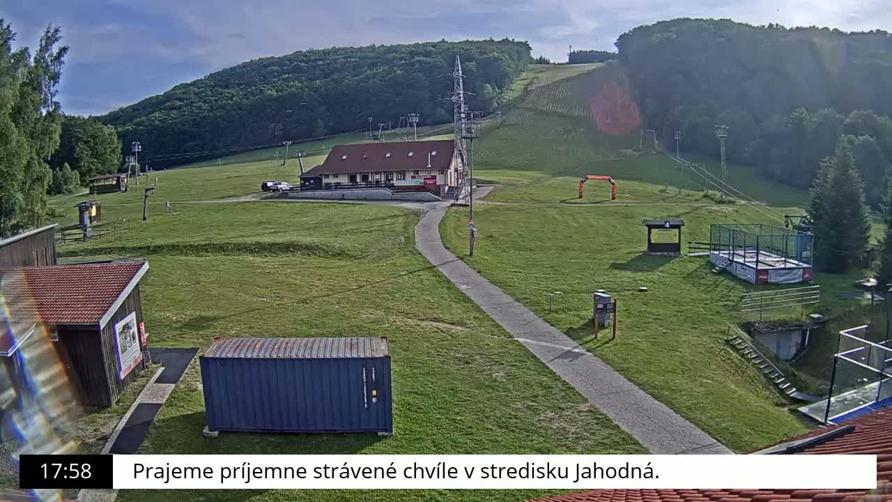 A grassy hill with a building, ski lift infrastructure, and a paved path is visible under sunny skies.