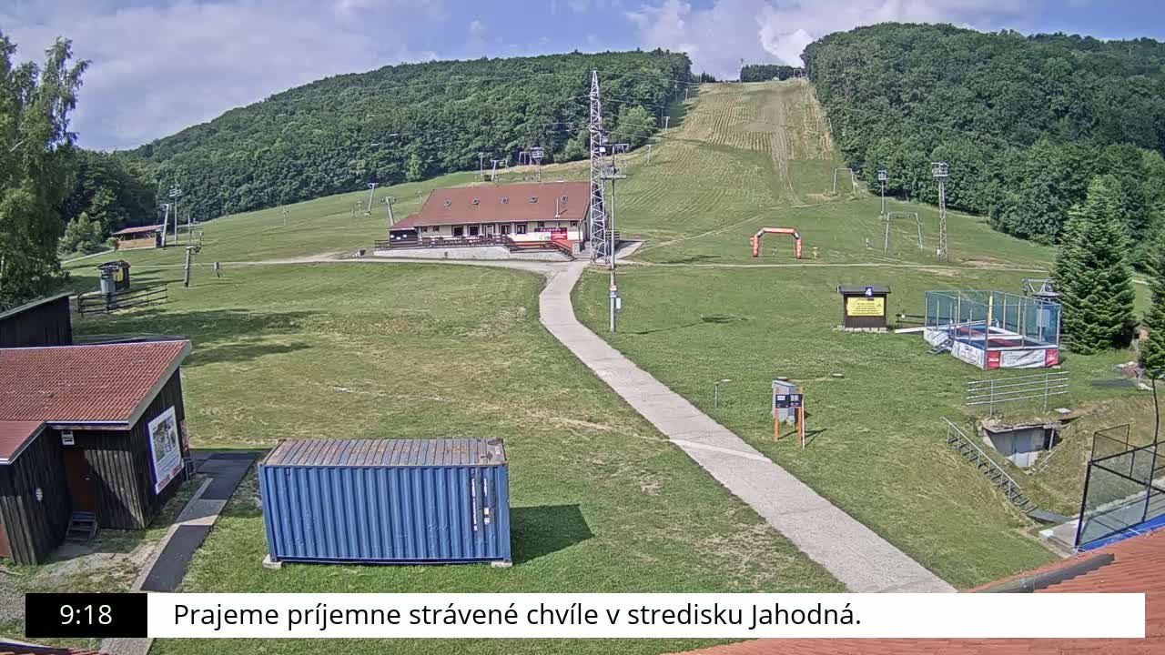 A grassy hillside with a ski lift, a building, and other recreational facilities is visible on a sunny day.