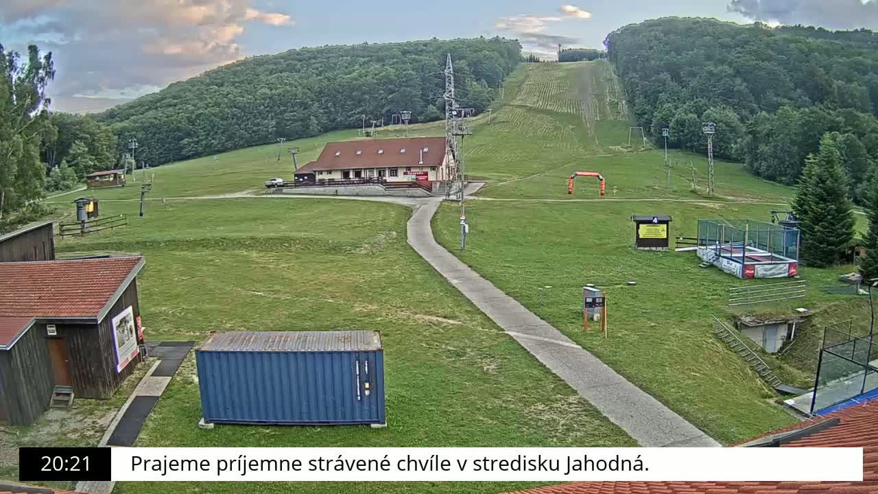 A grassy ski resort area with a building, lift infrastructure, and a trampoline under partly cloudy evening skies.