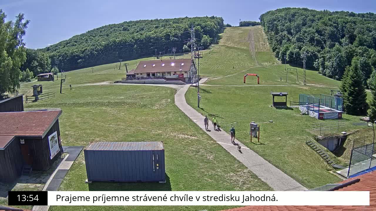 A sunny outdoor view of a grassy ski resort with several people walking a path near a building and a trampoline.