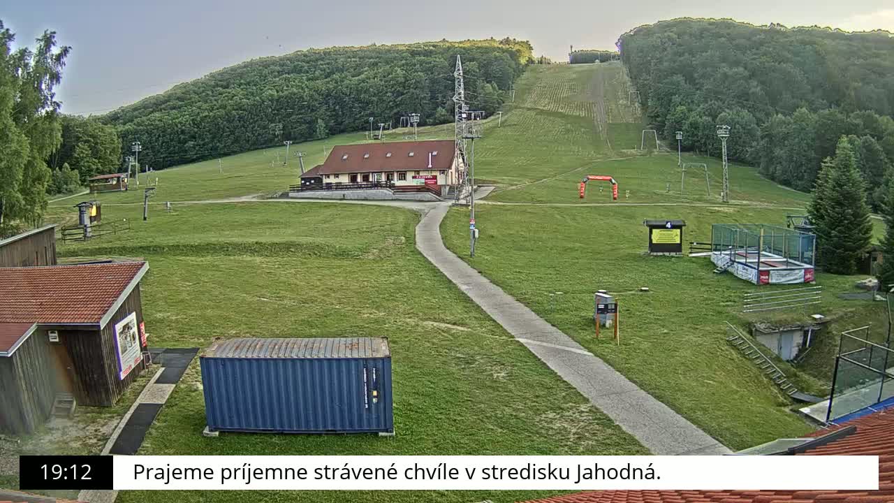 A grassy ski slope with a building, several smaller structures, and a paved pathway is visible under a mostly clear sky.