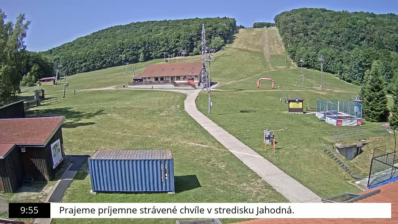 A grassy hillside with a ski lift, a building, and other recreational facilities is visible on a sunny day.