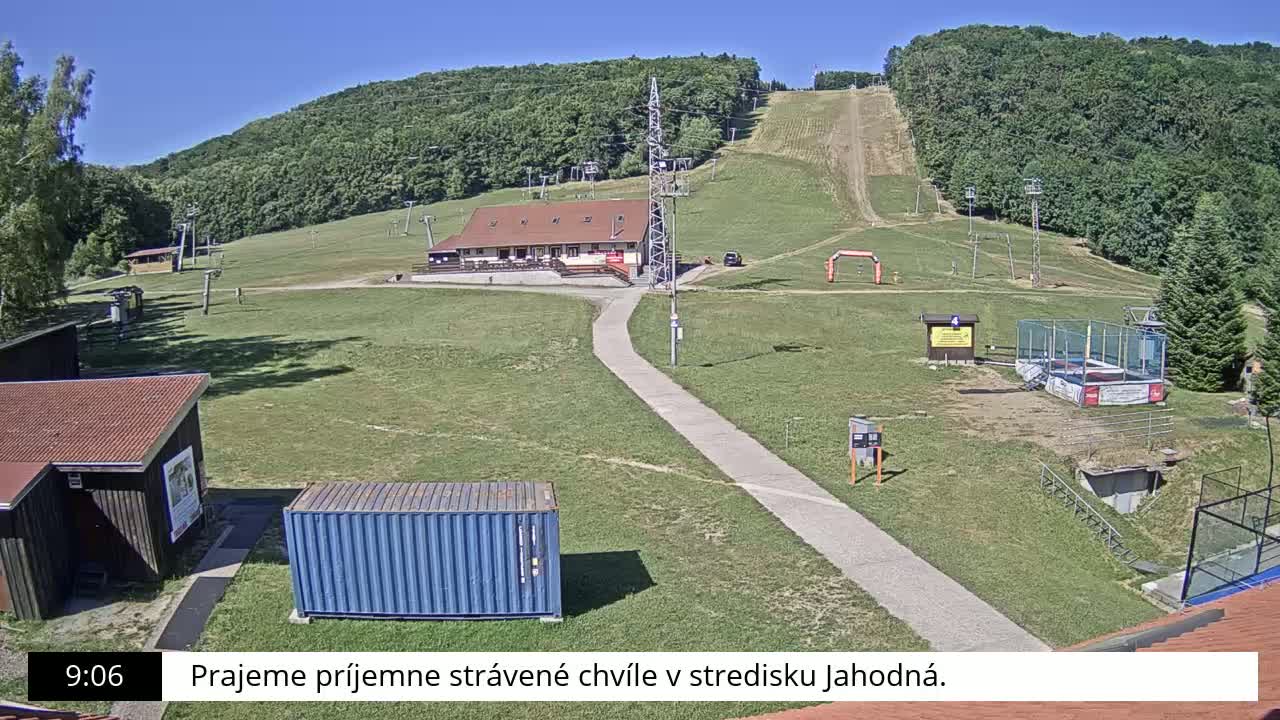 A ski resort on a sunny day, with a grassy area, several small buildings, a ski lift, and a paved pathway.