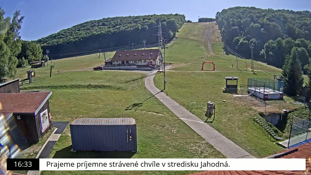 A grassy ski slope with a building, a paved path, and a shipping container is visible on a sunny day.