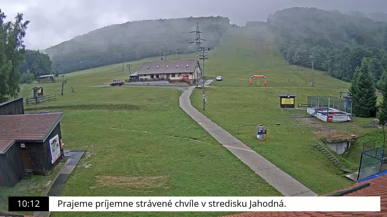 A grassy ski resort with a building, a paved path, and a foggy mountain in the background on an overcast day.