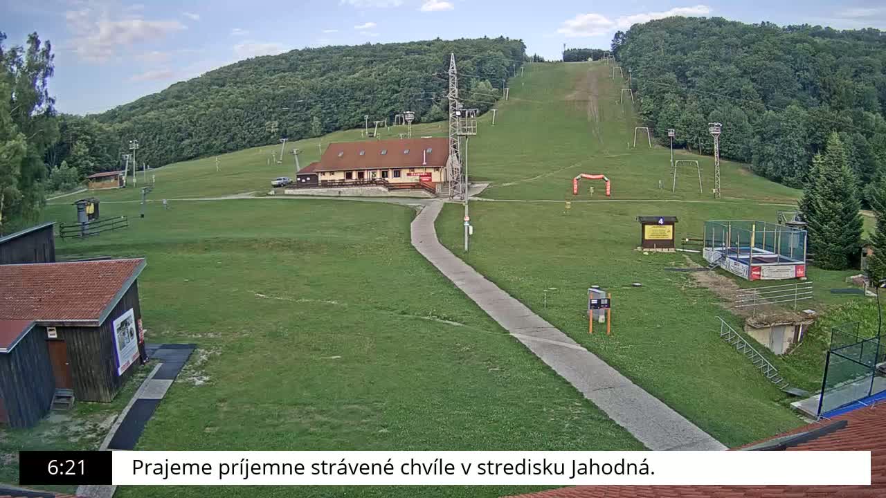 A grassy ski slope with a building, lift towers, and a few people walking on a paved path on a sunny day.