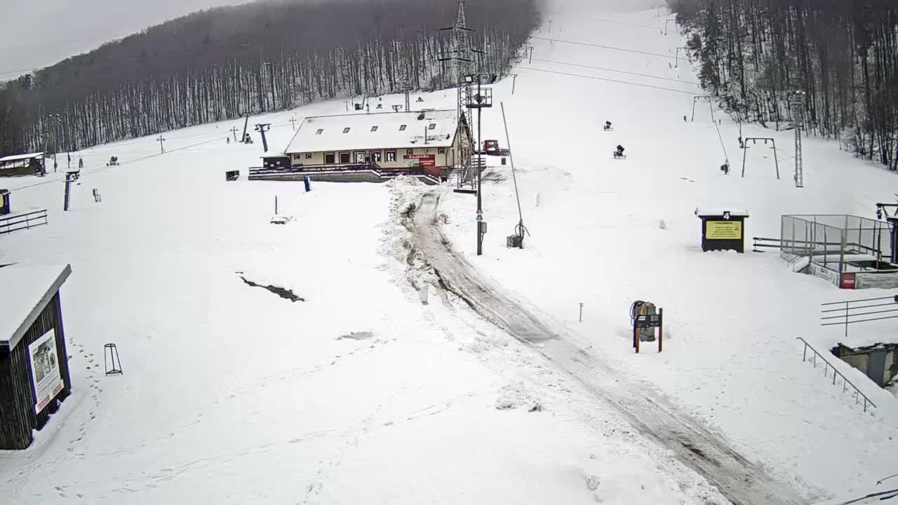 A snow-covered ski resort with various ski lifts, a central lodge, and a partially thawed access path is visible under an overcast sky.