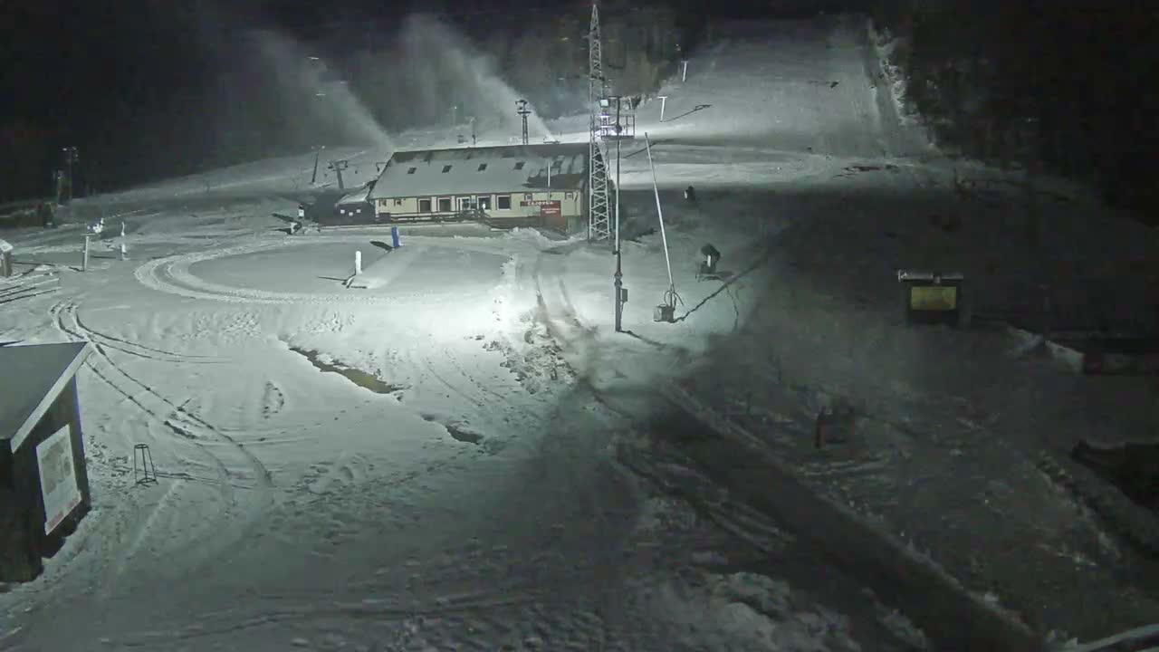 A snow-covered ski resort is depicted at night, illuminated by artificial lights, with multiple snow cannons actively blowing snow on the slopes under clear, cold conditions.