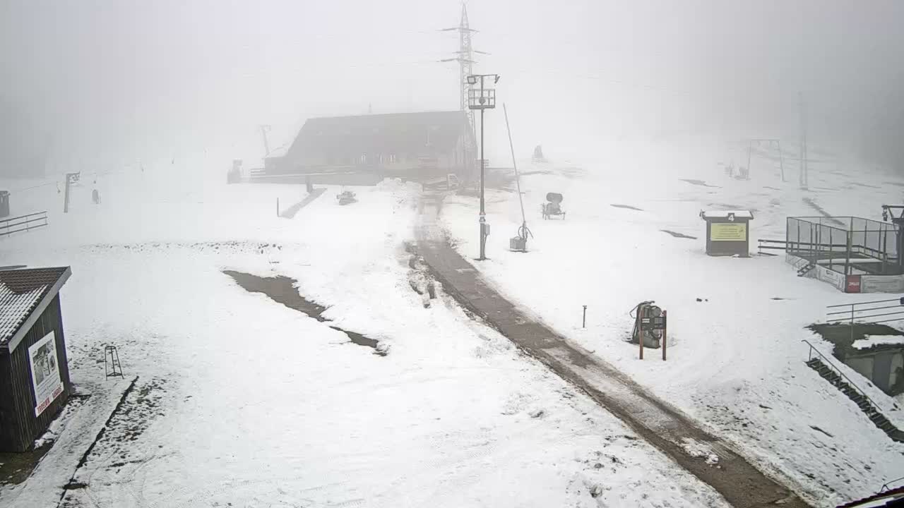 A snowy mountain landscape is shrouded in heavy fog, revealing a ski resort with several buildings, ski lift infrastructure, snow cannons, and a winding dirt path.