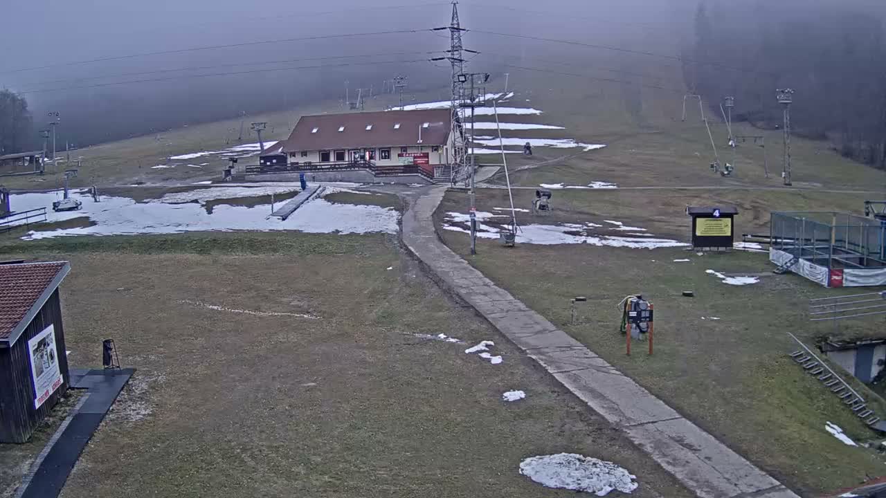 A snowy mountain landscape is shrouded in heavy fog, revealing a ski resort with several buildings, ski lift infrastructure, snow cannons, and a winding dirt path.