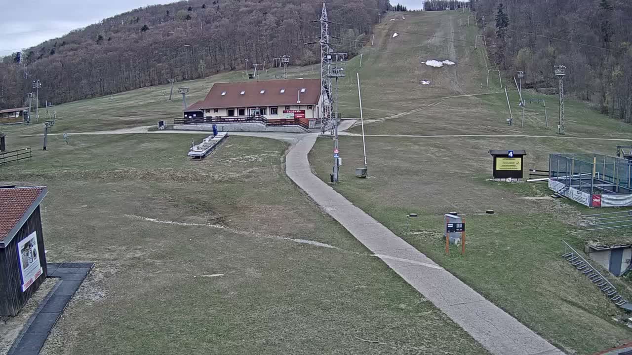 A snowy mountain landscape is shrouded in heavy fog, revealing a ski resort with several buildings, ski lift infrastructure, snow cannons, and a winding dirt path.