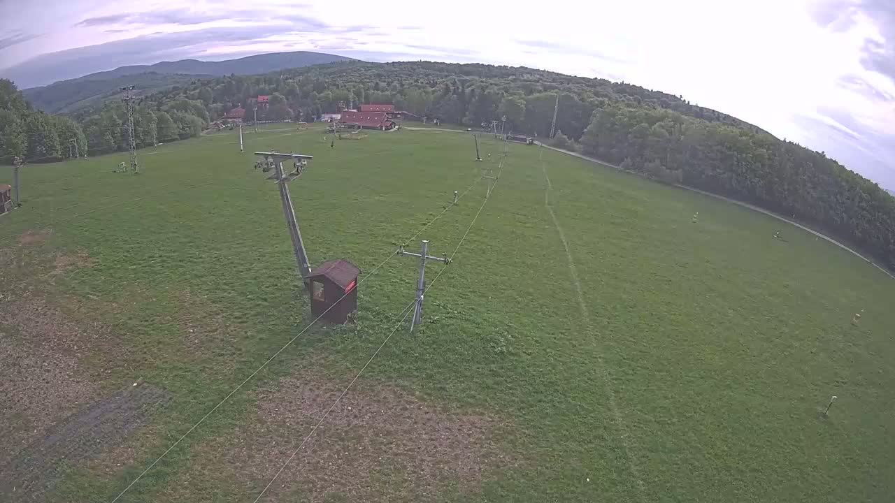 An aerial view shows a grassy field with a ski lift and a small building, under a partly cloudy sky, surrounded by a line of trees and distant mountains.