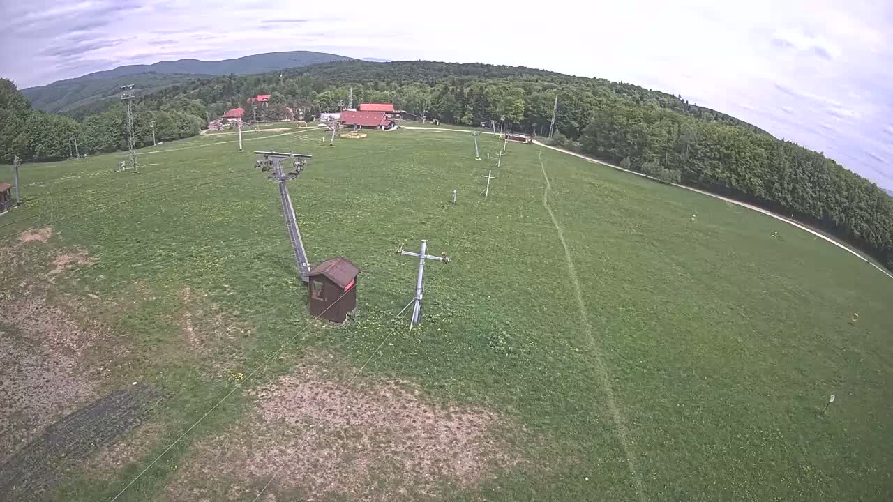 An aerial view shows a grassy field with a ski lift and support structures, under a partly cloudy sky, surrounded by a forest and distant mountains.