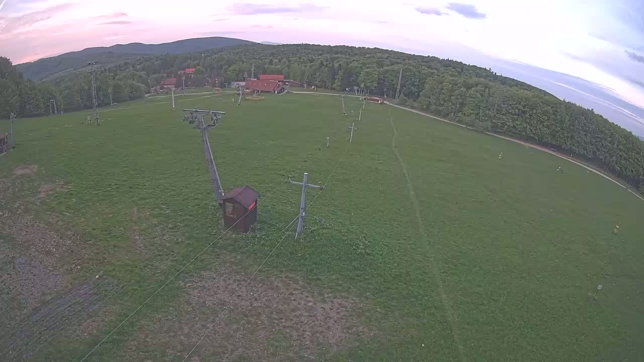 An aerial view shows a grassy field with a ski lift and small buildings, surrounded by a forest under a partly cloudy sky.