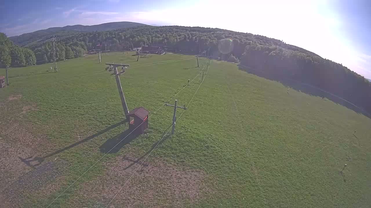 An aerial view shows a grassy field with a ski lift in the foreground, backed by a line of trees and distant mountains under a mostly sunny sky.