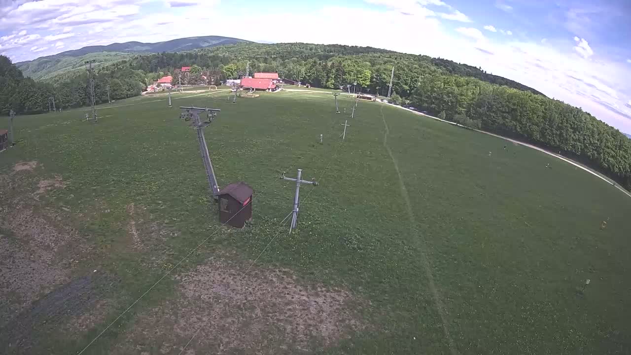 An aerial view shows a grassy field with a chairlift and supporting structures, bordered by a forest under a partly cloudy sky.