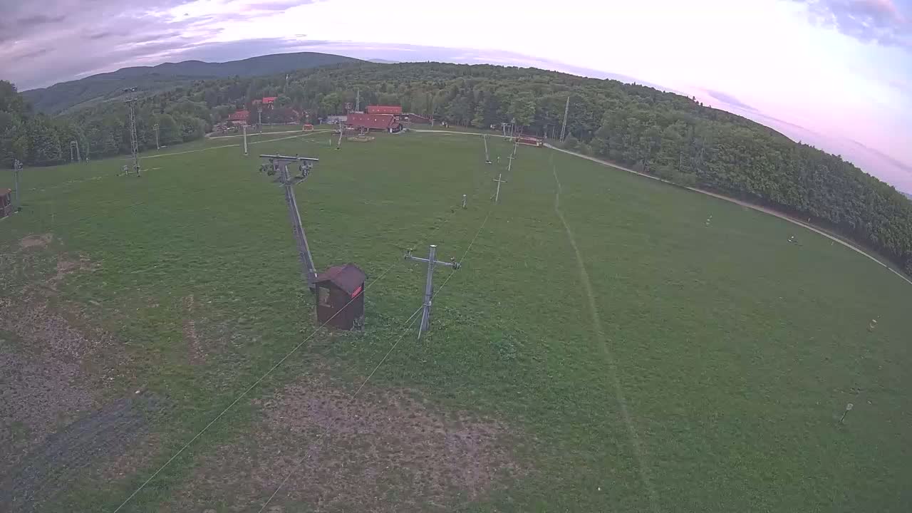 An aerial view shows a grassy field with a ski lift and a few buildings in the distance under a cloudy sky.