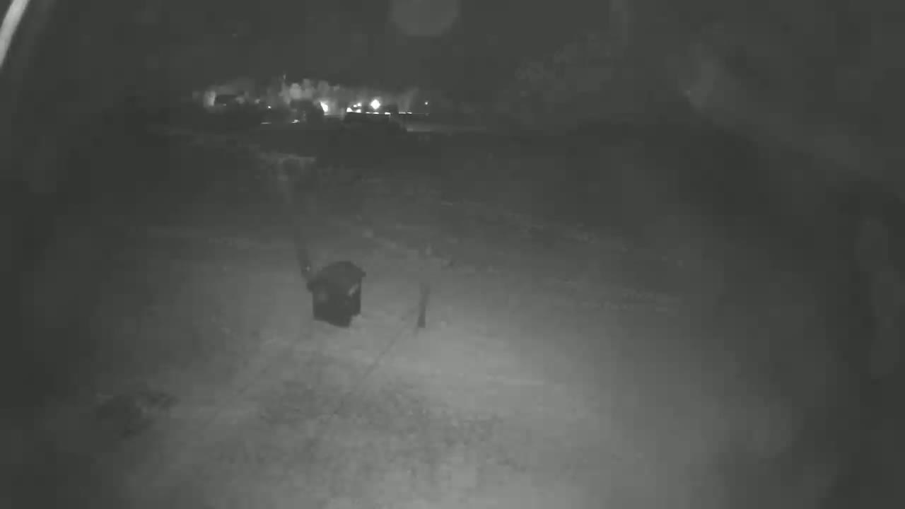 A nighttime, low-light image shows a dark, possibly trash, container in a snow-covered area.