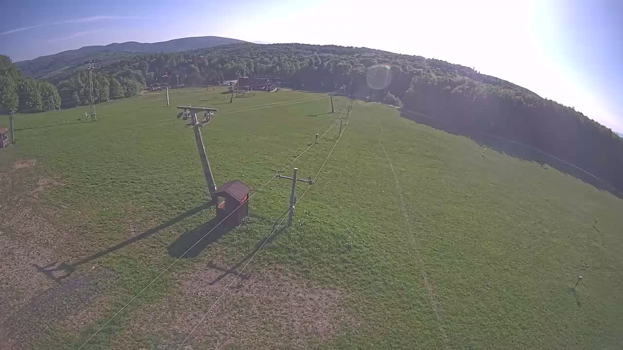 A grassy field with a ski lift and small structure is visible under a sunny sky, backed by a line of trees and distant mountains.
