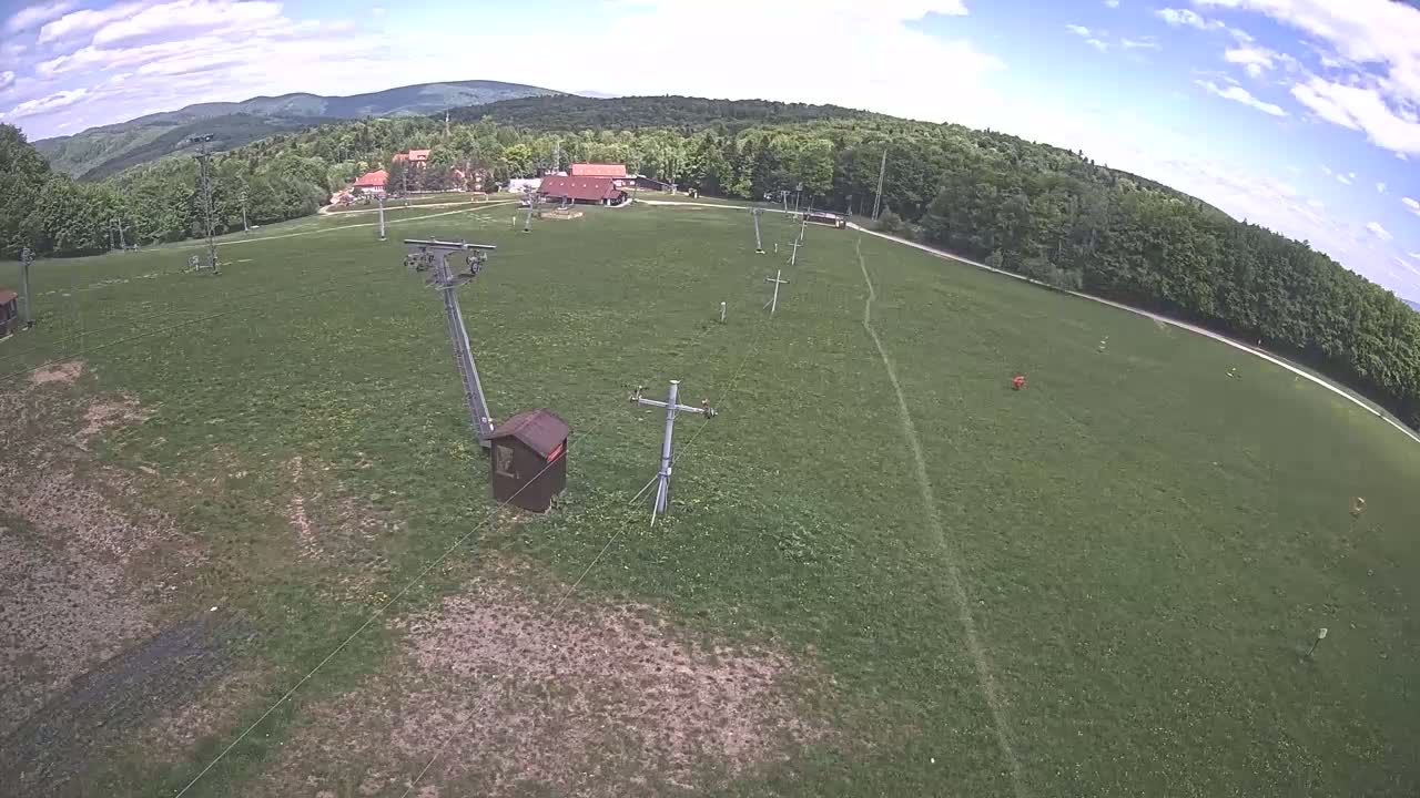 An aerial view shows a grassy field with a chairlift system, a few small buildings, and a forest under a partly cloudy sky.