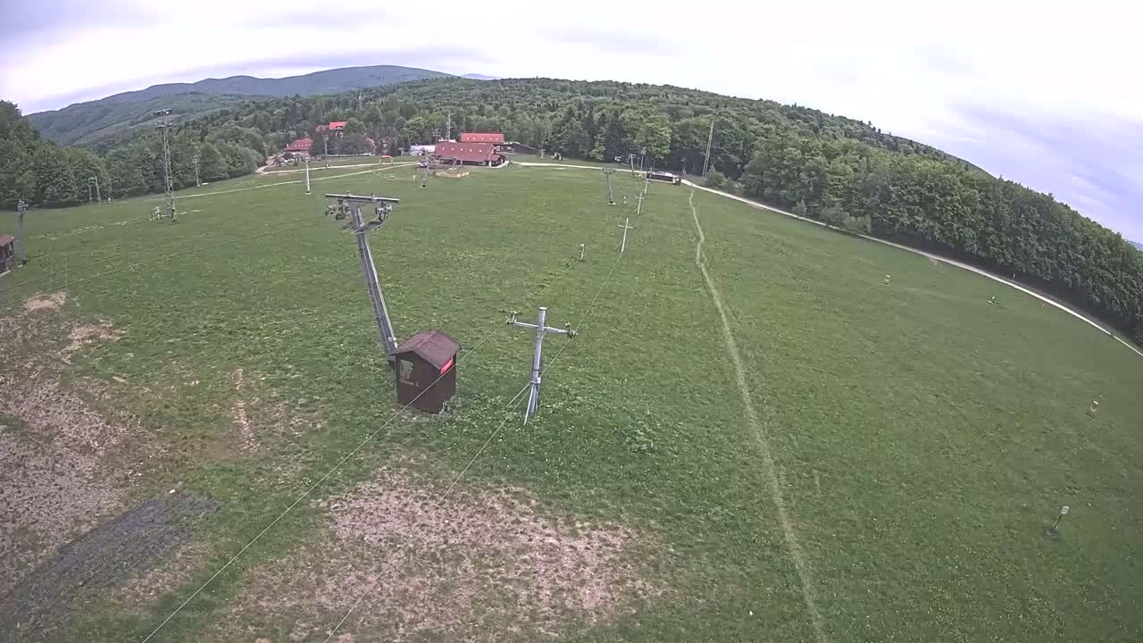An overhead view shows a grassy field with a chairlift infrastructure, a few buildings in the distance, and a line of trees on a cloudy day.