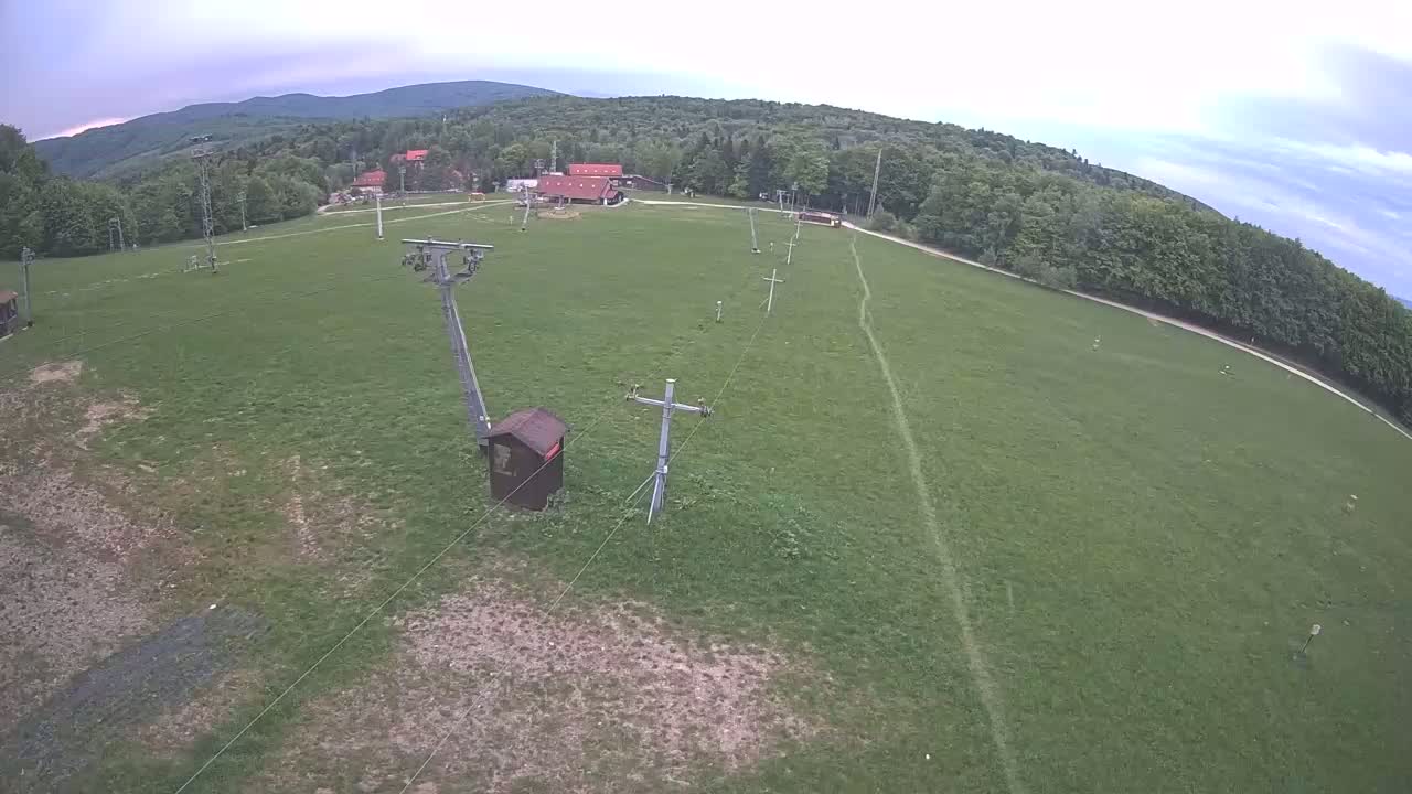 An aerial view shows a grassy field with a ski lift and small buildings, surrounded by a forest under a cloudy sky.