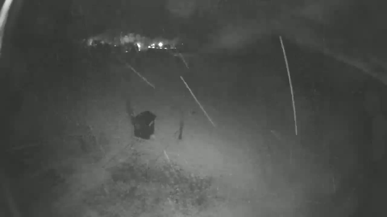 A dark, nighttime outdoor scene shows a blurry figure near the bottom center of the frame, with several streaks of light appearing to fall from above in snowy or rainy conditions.