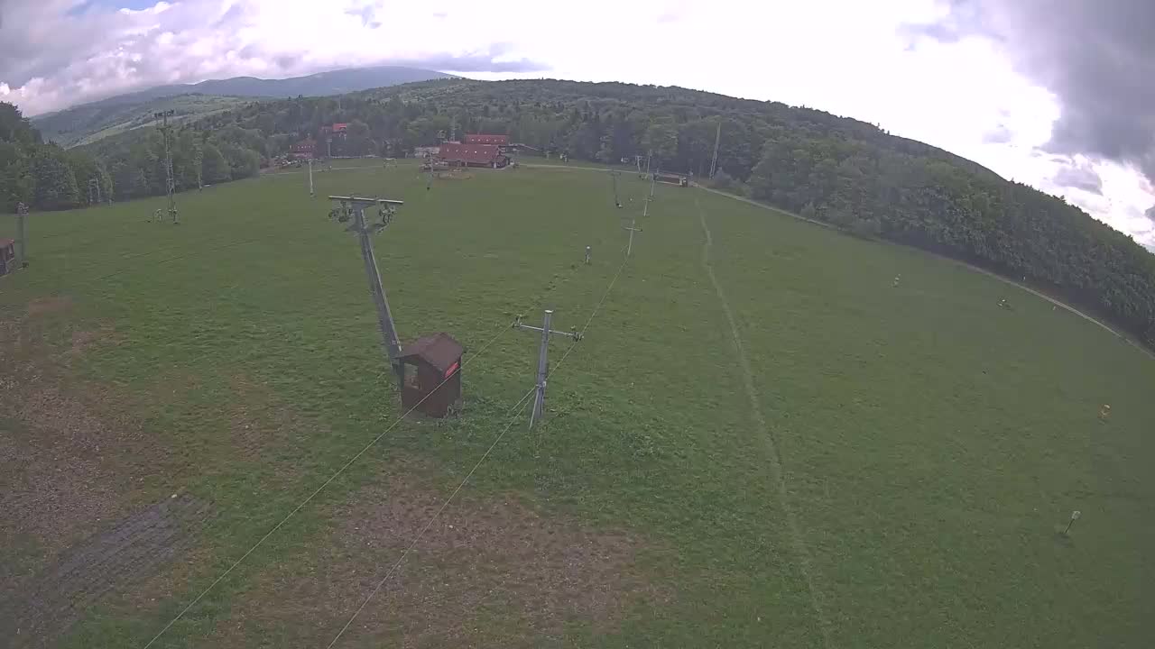 An overhead view shows a grassy field with a ski lift and a few buildings in the background under a partly cloudy sky.